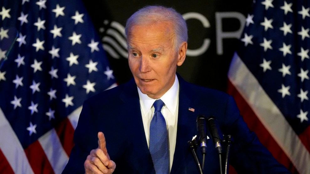 Joe Biden onstage in a navy suit, pointing with one of his fingers. Two American flags stand behind him.