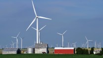 Dozens of wind turbines tower over a rural field with a silo and other farm buildings.