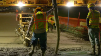Two workers wearing neon vests watch concrete being poured.