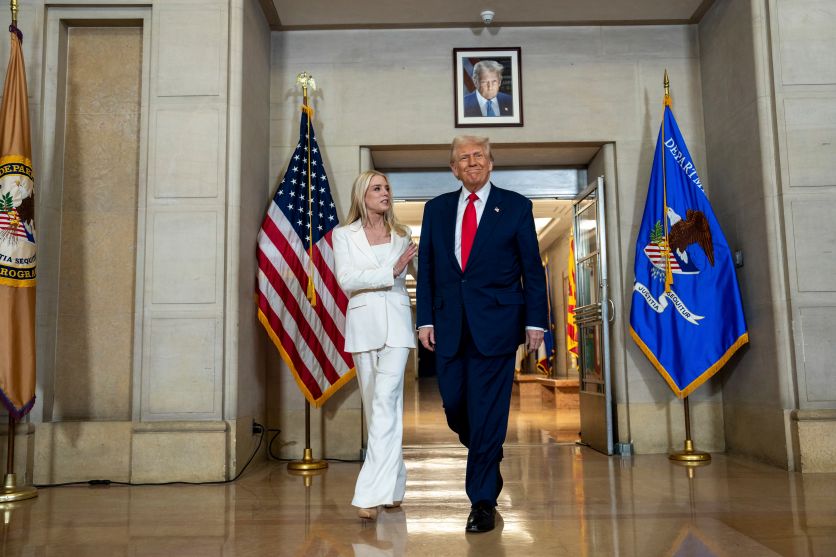 Pam Bondi, dressed in an all-white suit, and Donald Trump, dressed in a navy suit and red tie, walk into a large room with smooth stone walls.