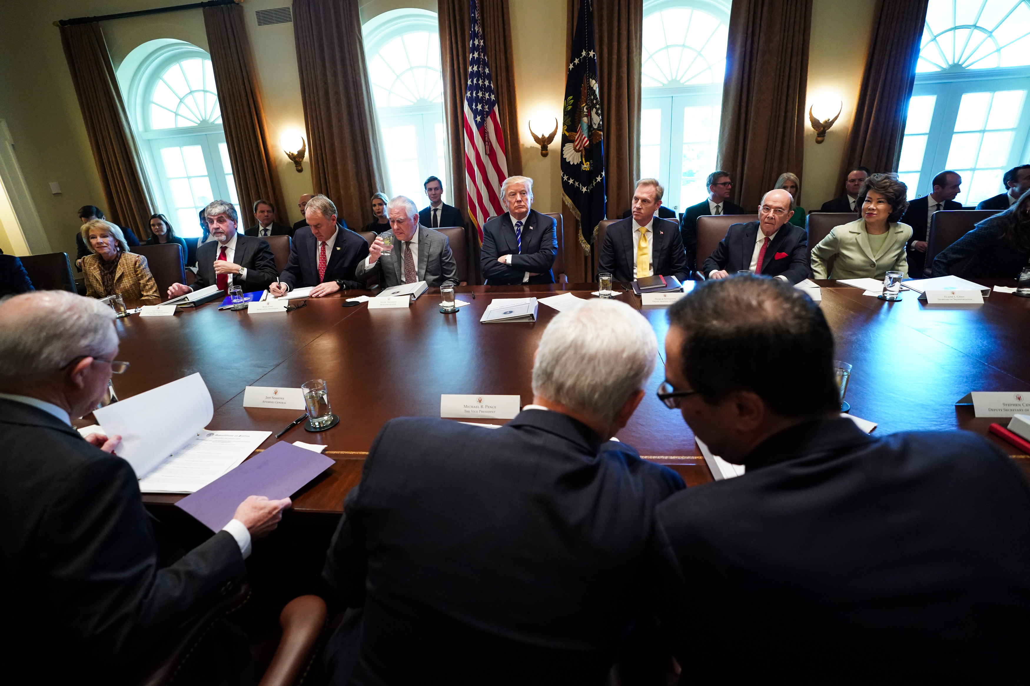 President Trump sitting with his arms crossed at a large table surrounded by Cabinet members.