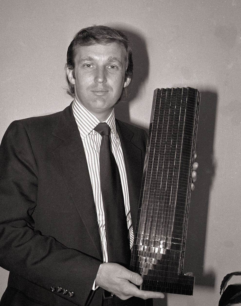 A black and white photo of a young Donald Trump wearing a suit and holding a model of a tall building.