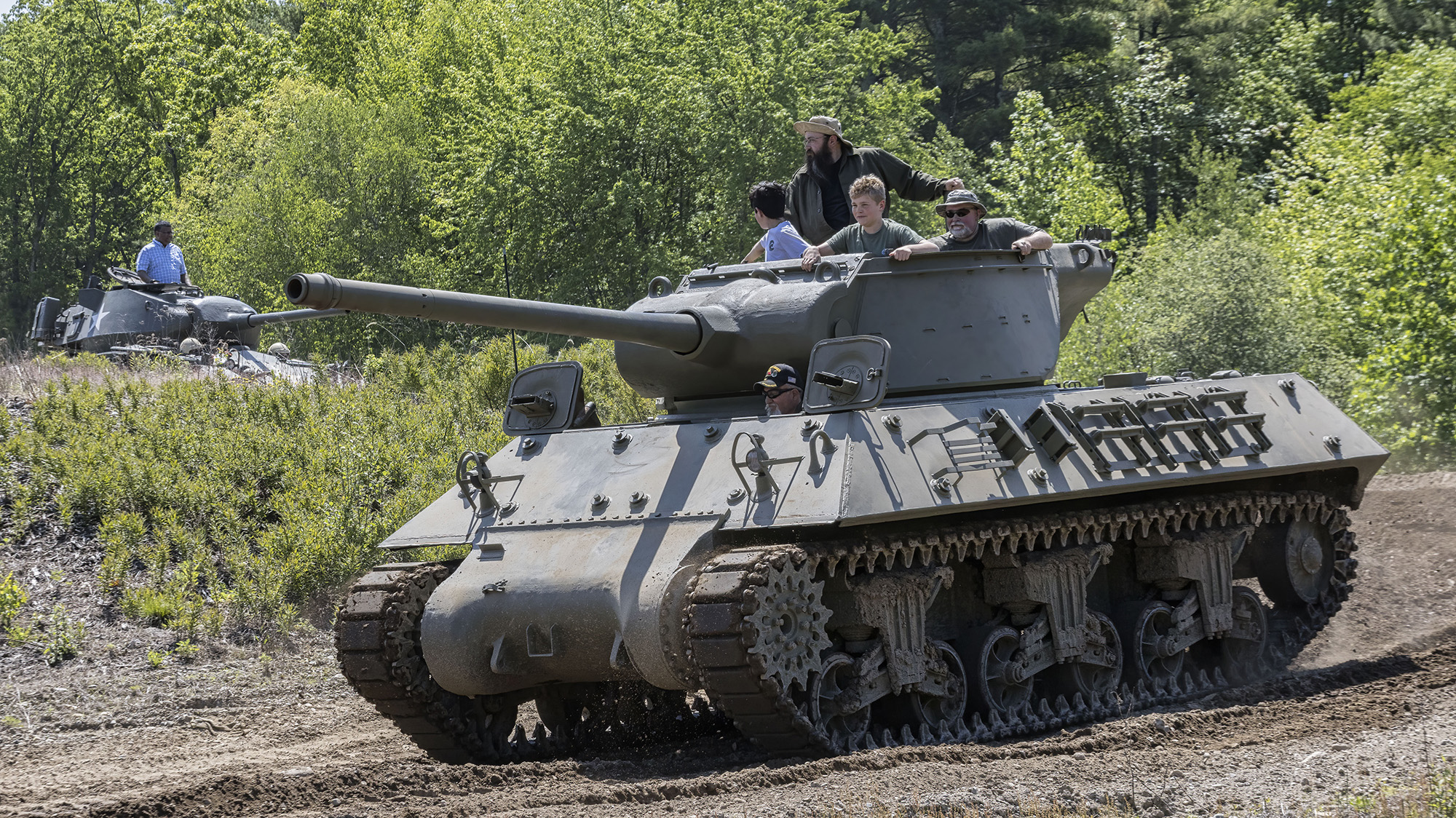 A tank with four people sitting in the top turret. In the background, a second tank has one person standing in the top turret.