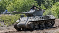 A tank with four people sitting in the top turret. In the background, a second tank has one person standing in the top turret.