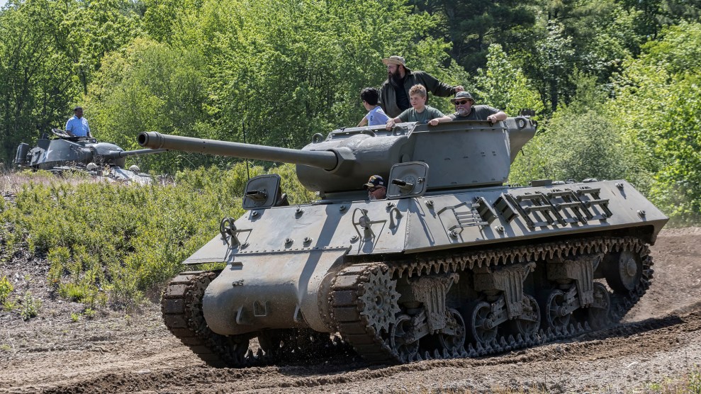A tank with four people sitting in the top turret. In the background, a second tank has one person standing in the top turret.