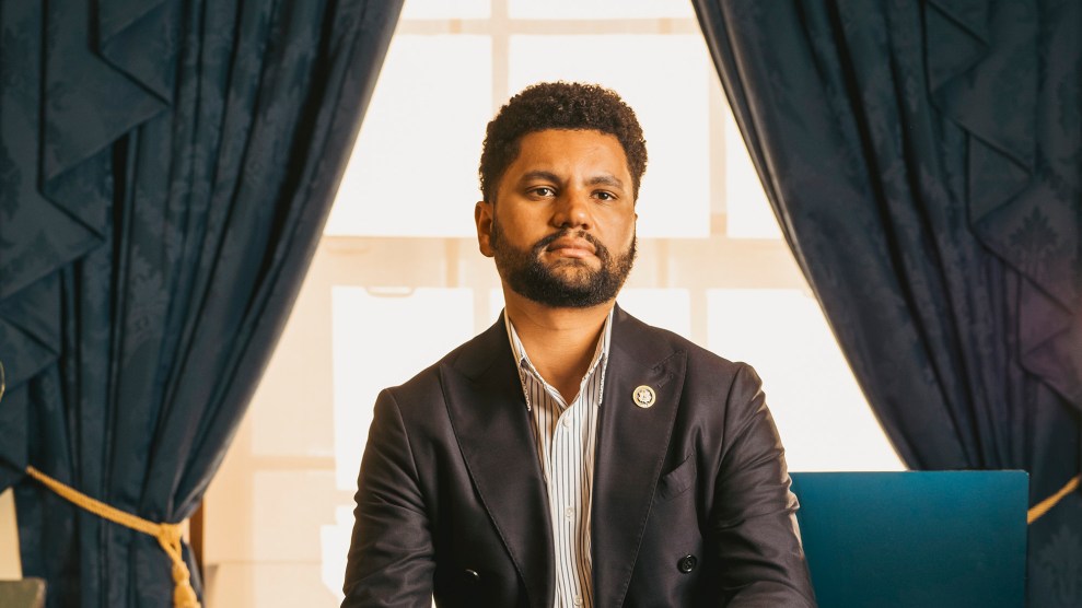 Representative Maxwell Frost sits on his desk in his office in D.C.; his expression is serious and his gaze looks off beyond the viewer.