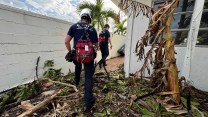 Two people wearing white hard hats and red backpacks walk through a yard full of debris.