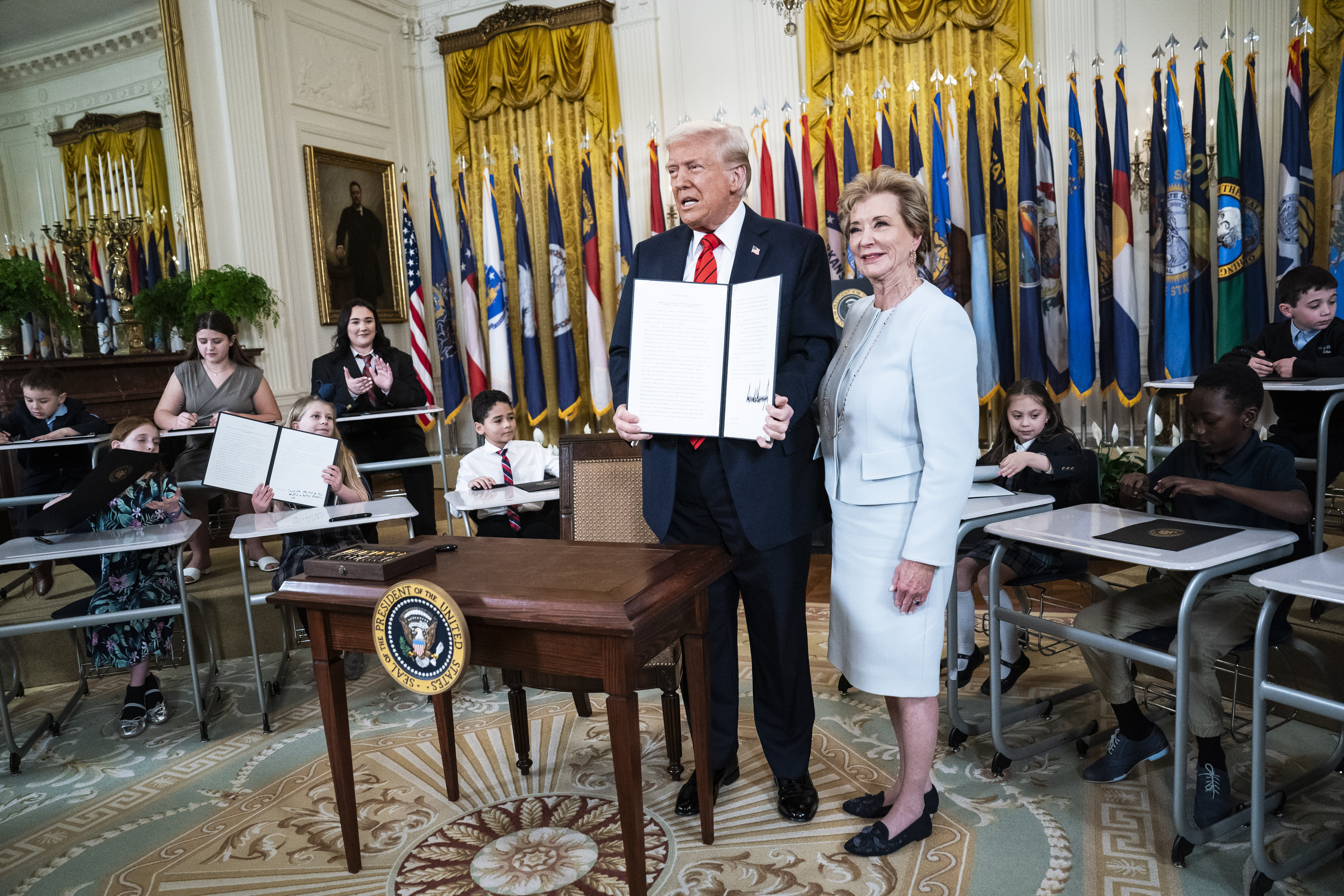 Donald Trump standing next to Linda McMahon, holding a signed order, surrounded by school children at desks.