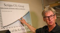 A white man with grey hair points at a diagram showing a blue line curving upward.
