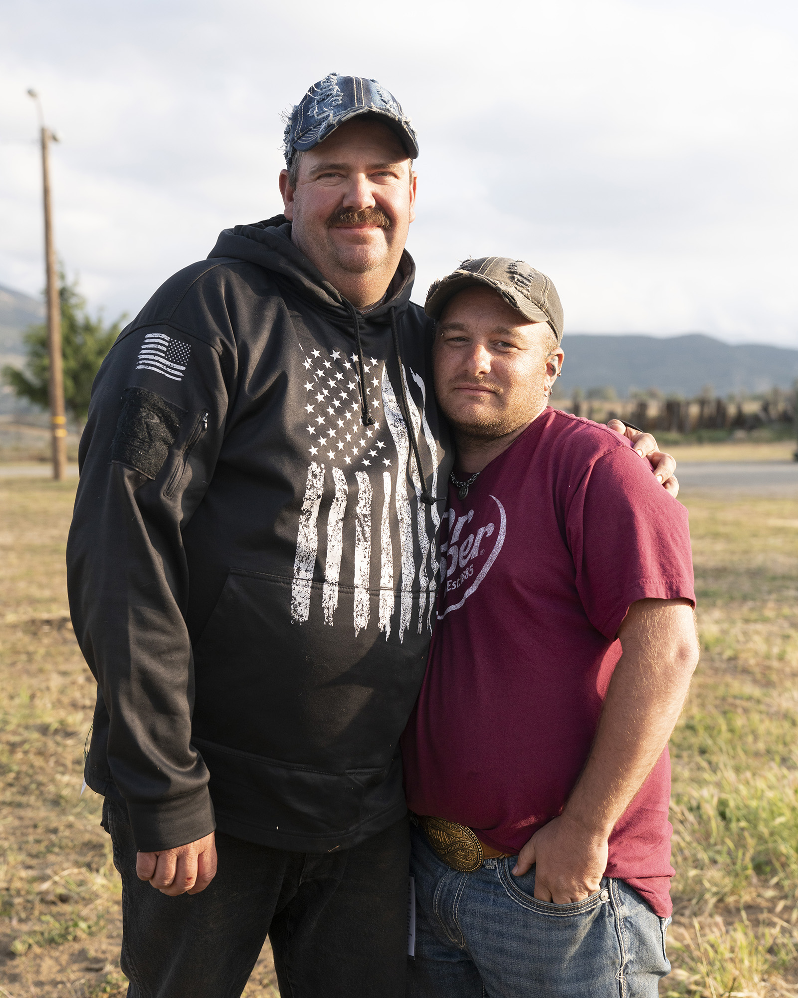 Portrait of two men wearing baseball hats standing in a field, with an arm around each other.
