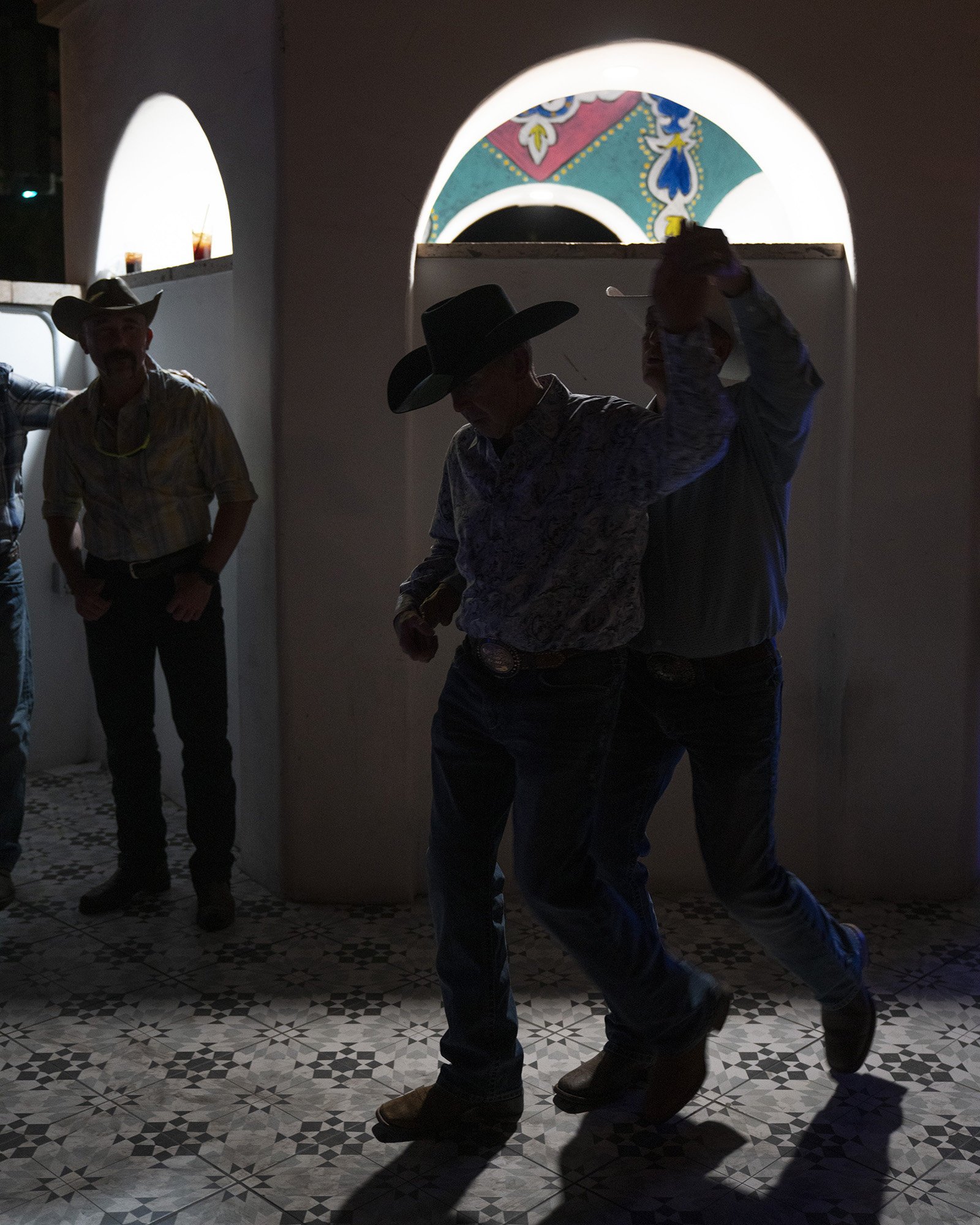 Two men in cowboy hats dancing in a dimly lit room with a mosaic tile floor.