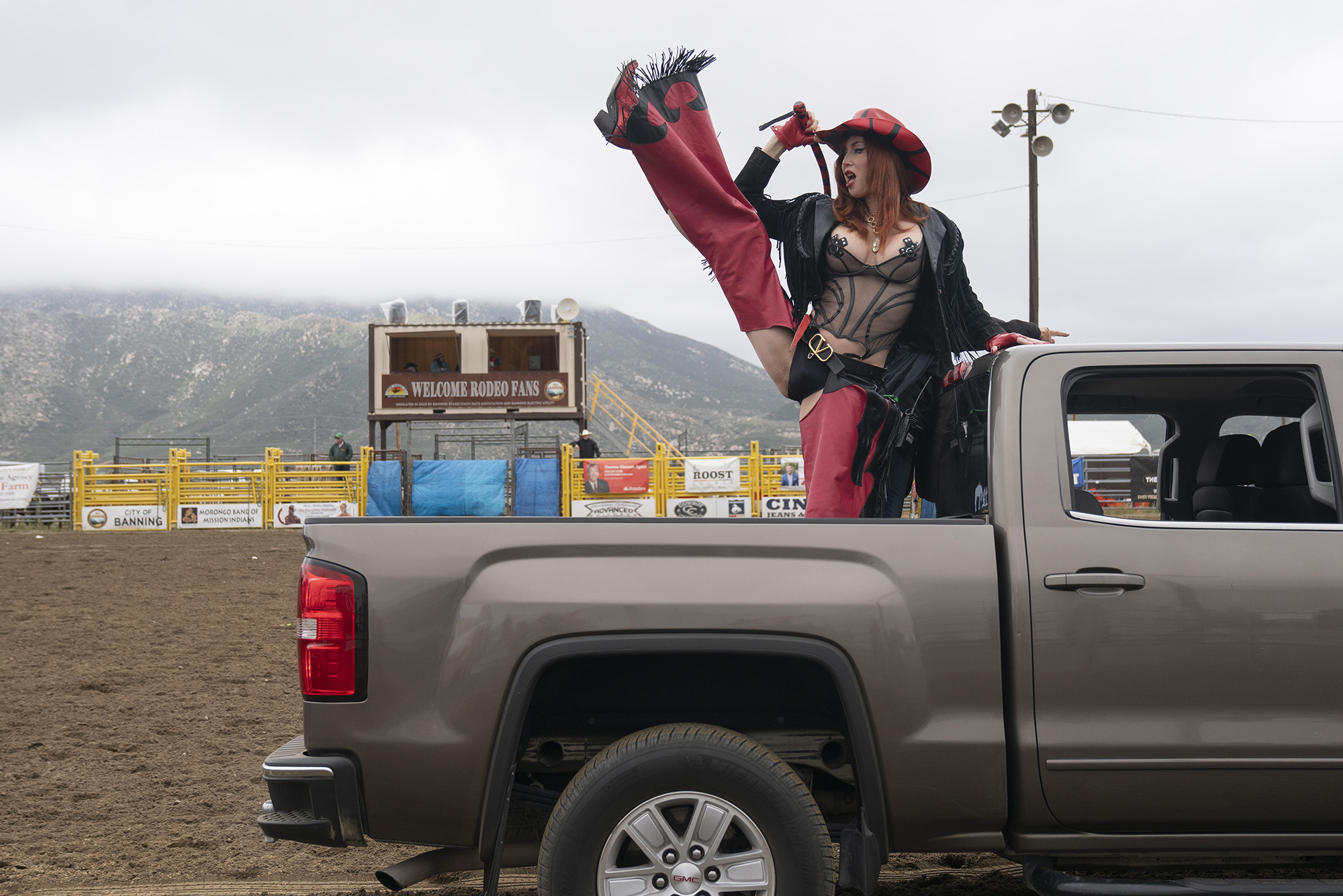 Person in the bed of a pickup truck performs a high kick. They wear a red cowboy hat, black lingerie-style corset, black jacket, and red-and-black chaps.