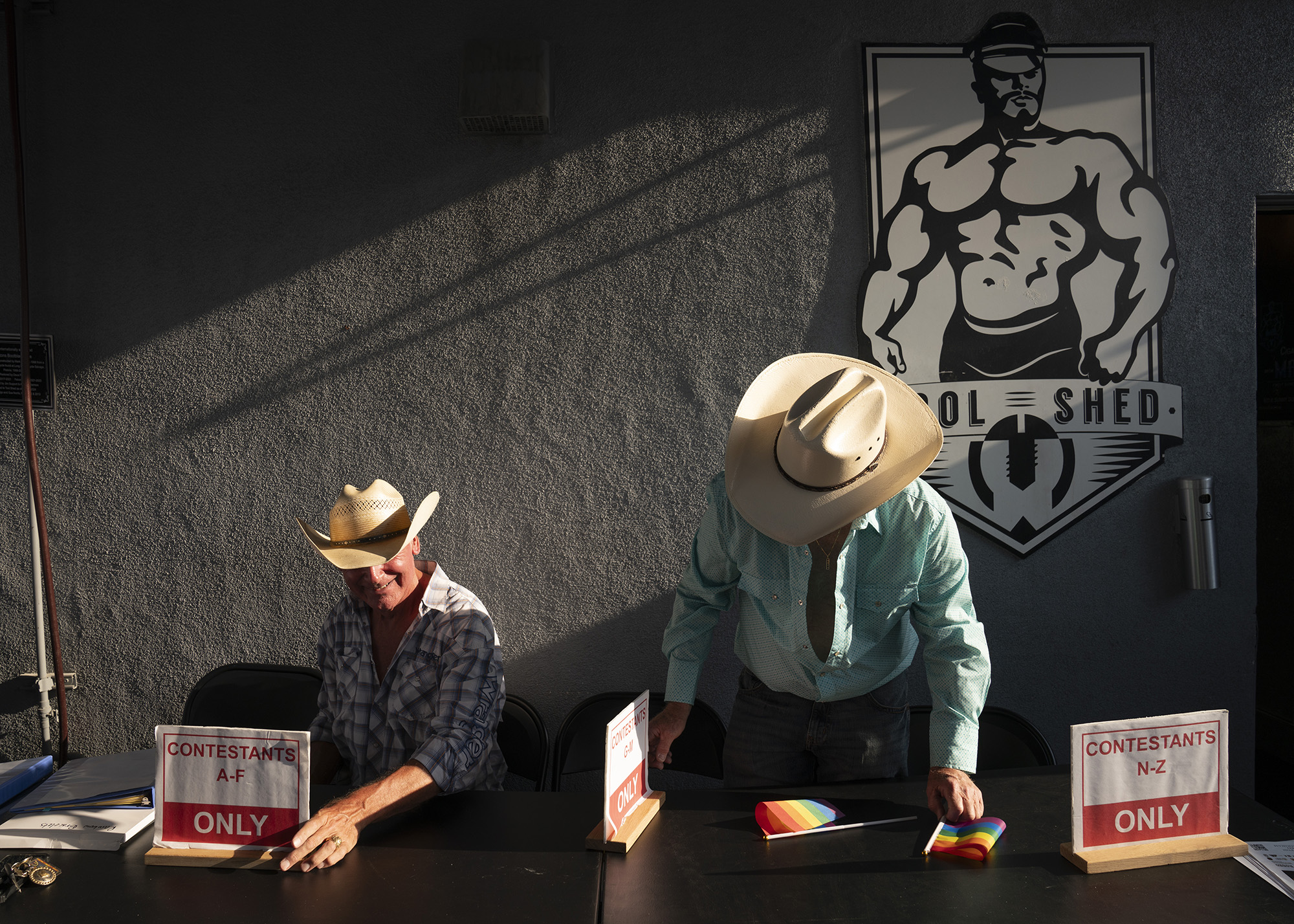 Two men at a desk wearing beige cowboy hats. Behind them is a sign for the Tool Shed, with a logo of a muscular shirtless man.