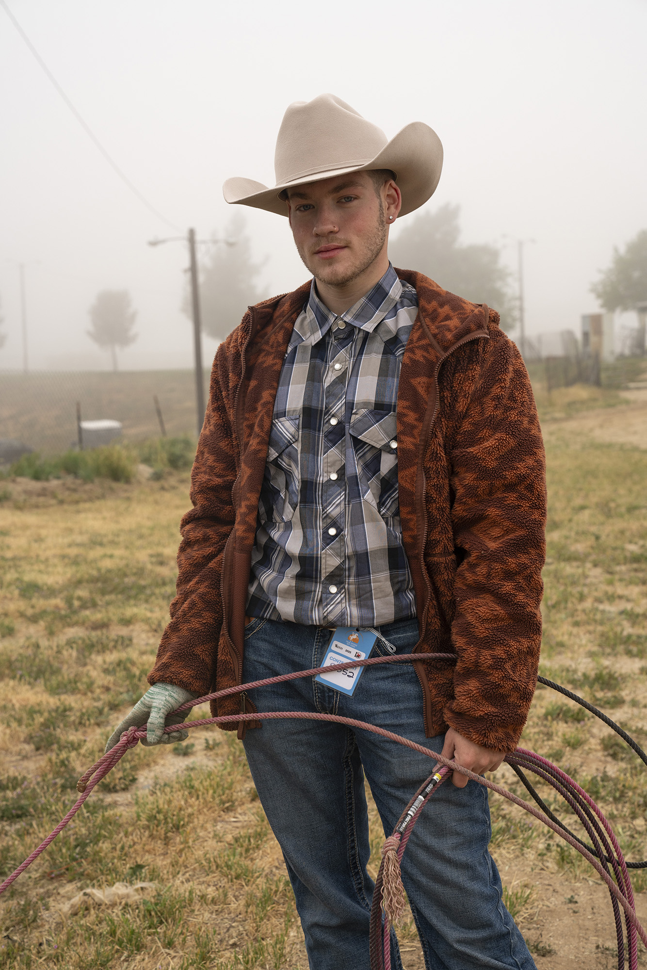Portrait of a person in tan cowboy hat, blue plaid shirt, and brown fleece jacket, holding a lasso.