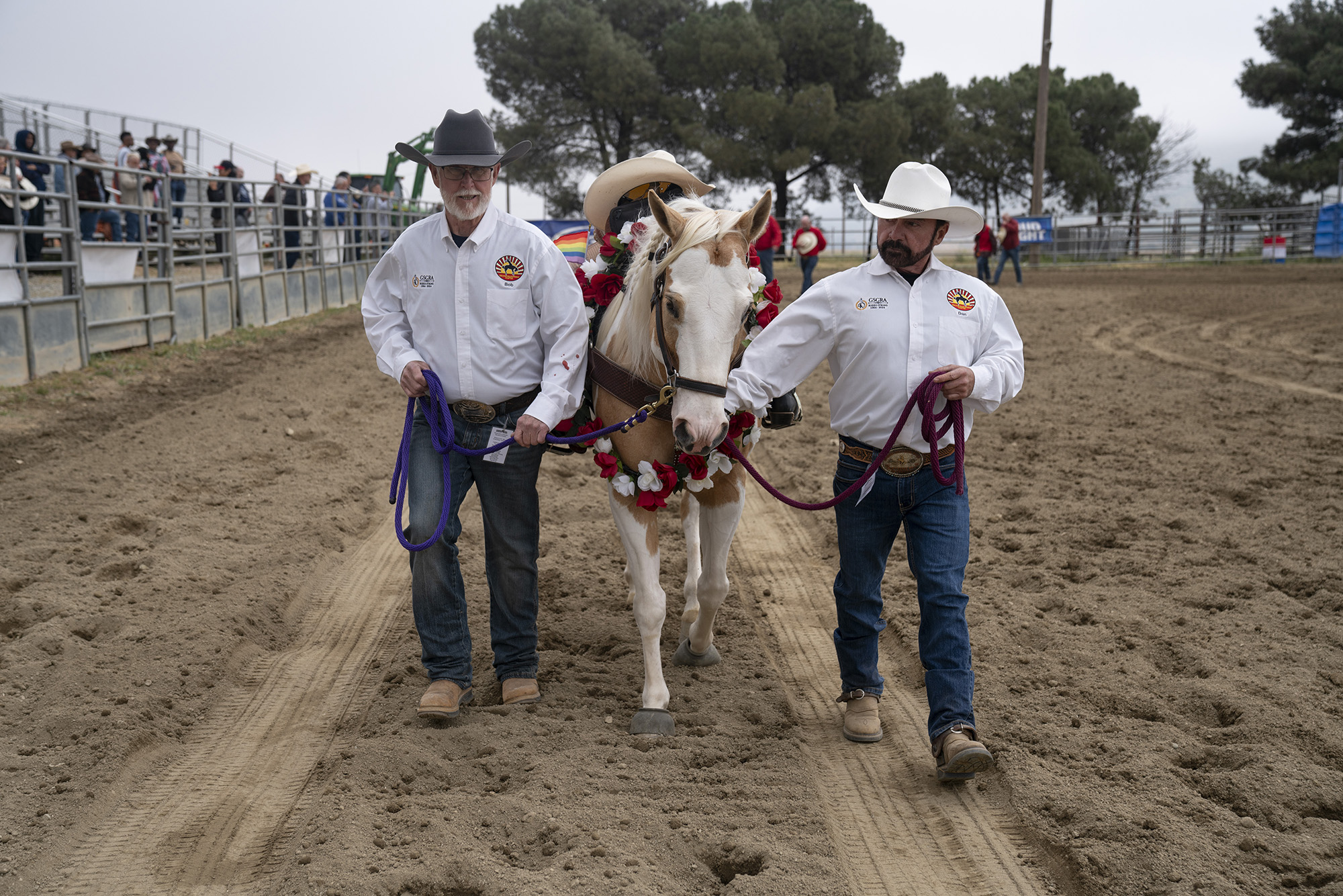Two men leading a horse around a dirt arena. The men wear matching white button-down shirts, jeans, and tan boots, with one man wearing a black cowboy hat and the other wearing a white one. The horse is draped with a red-and-white floral wreath with a beige cowboy hat atop its saddle.