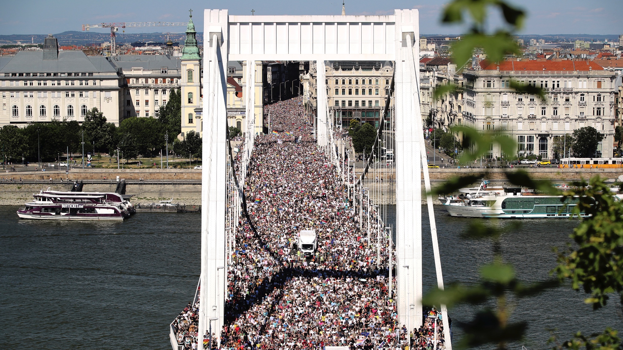 People walking fill the deck of a multilane, white suspension bridge