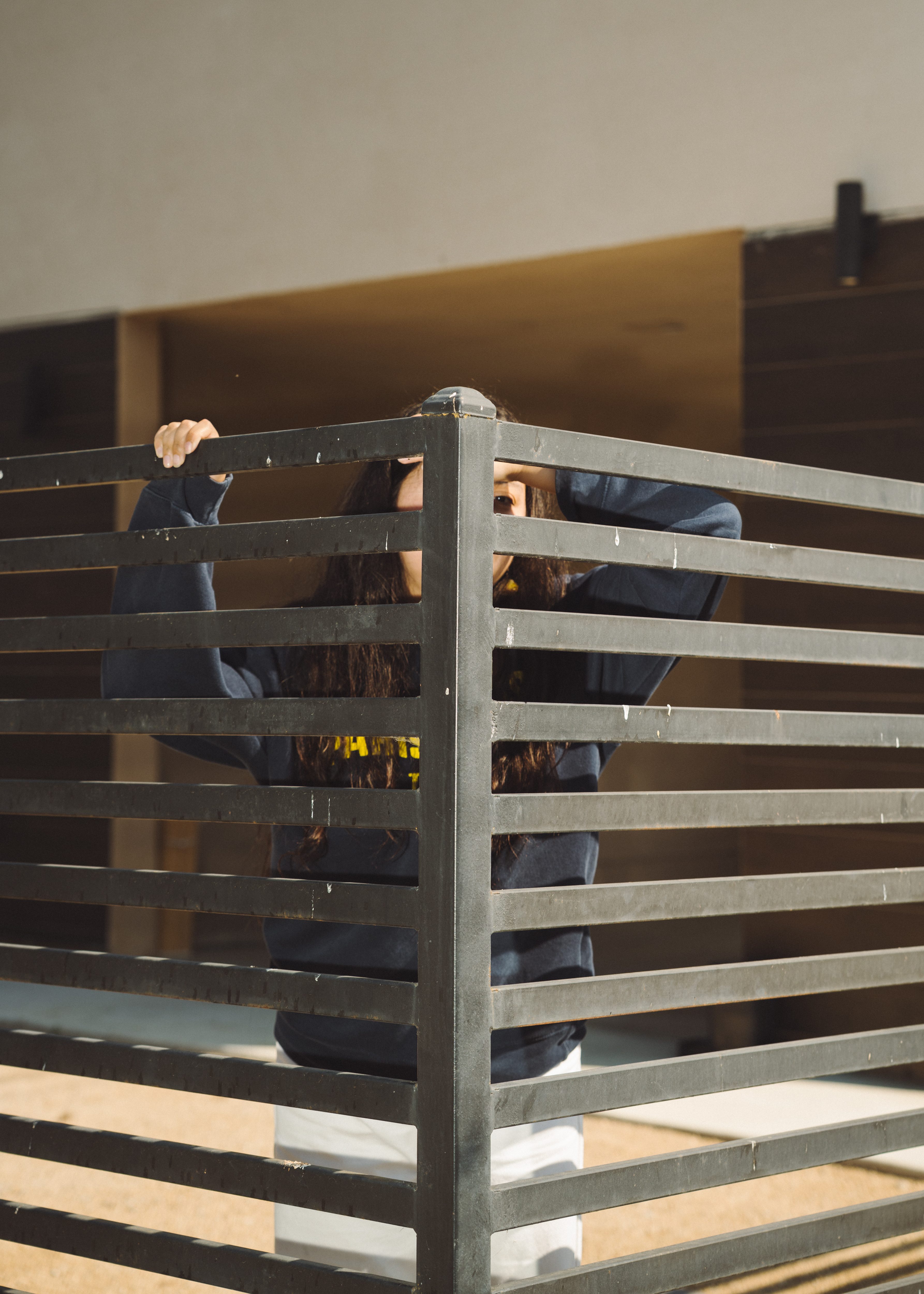 A young woman stands behind a slatted iron fence, her face obscured by horizontal beams.