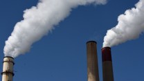 Close-up of the tops of three smokestacks, two with billowing white smoke against a bright blue sky.