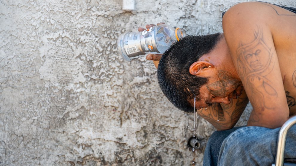 A shirtless Asian man with tattoos sits in a chair next to a stucco wall, looking miserable and sweltering, with a water bottle in his hand.