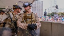A Marine man in uniform looks away from the protestors in a city. Another looks towards them.