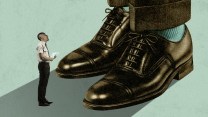 A small man in a shirt and tie holds tax forms as he looks up at the gigantic shoes, feet, and lower legs of another man.