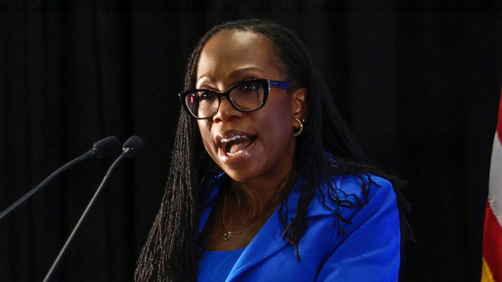 Ketanji Brown Jackson stands behind two microphones while wearing a blue suit. Part of a US flag is visible behind her.