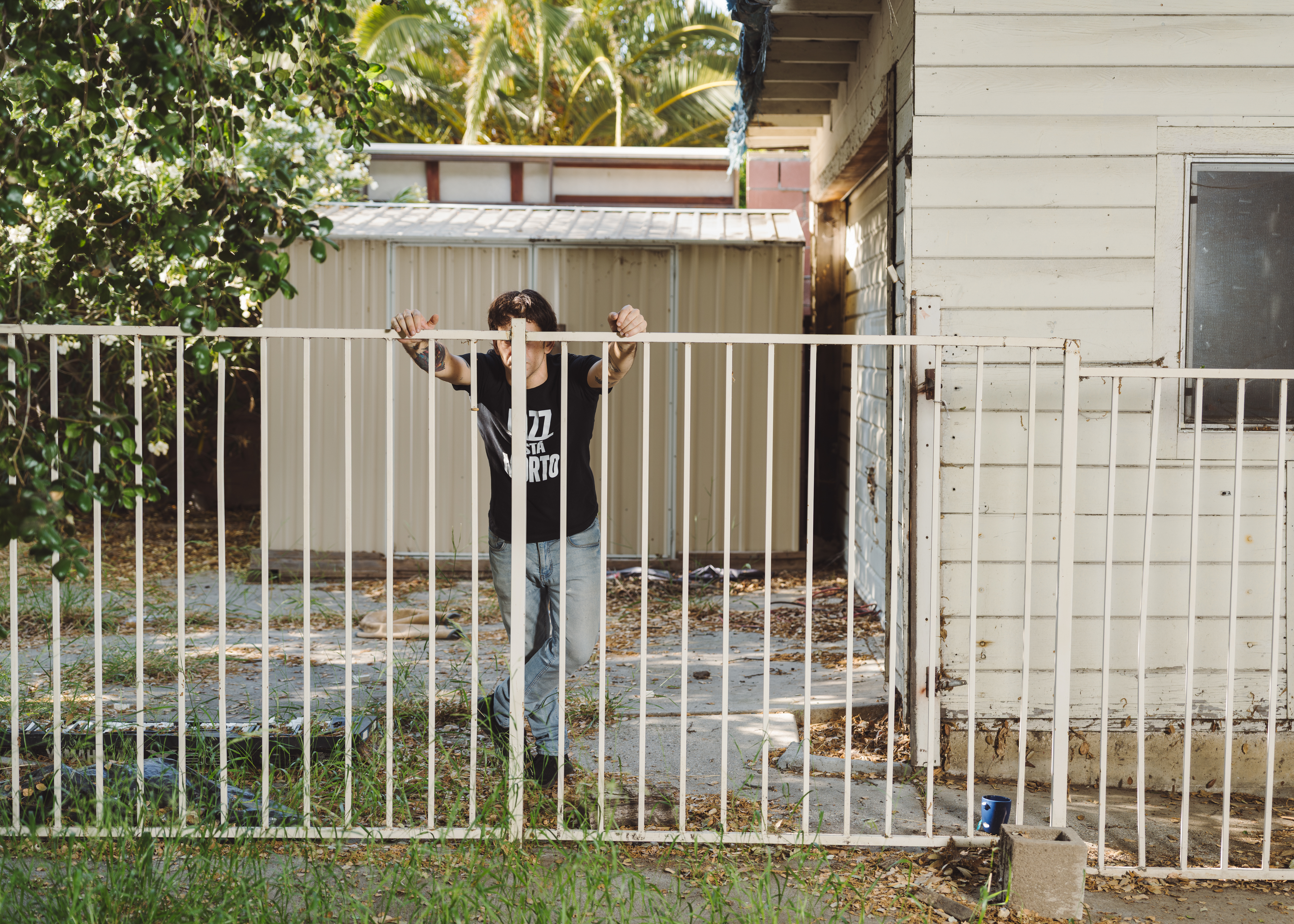 A man stands behind a white metal fence, his face obscured by the vertical slats.