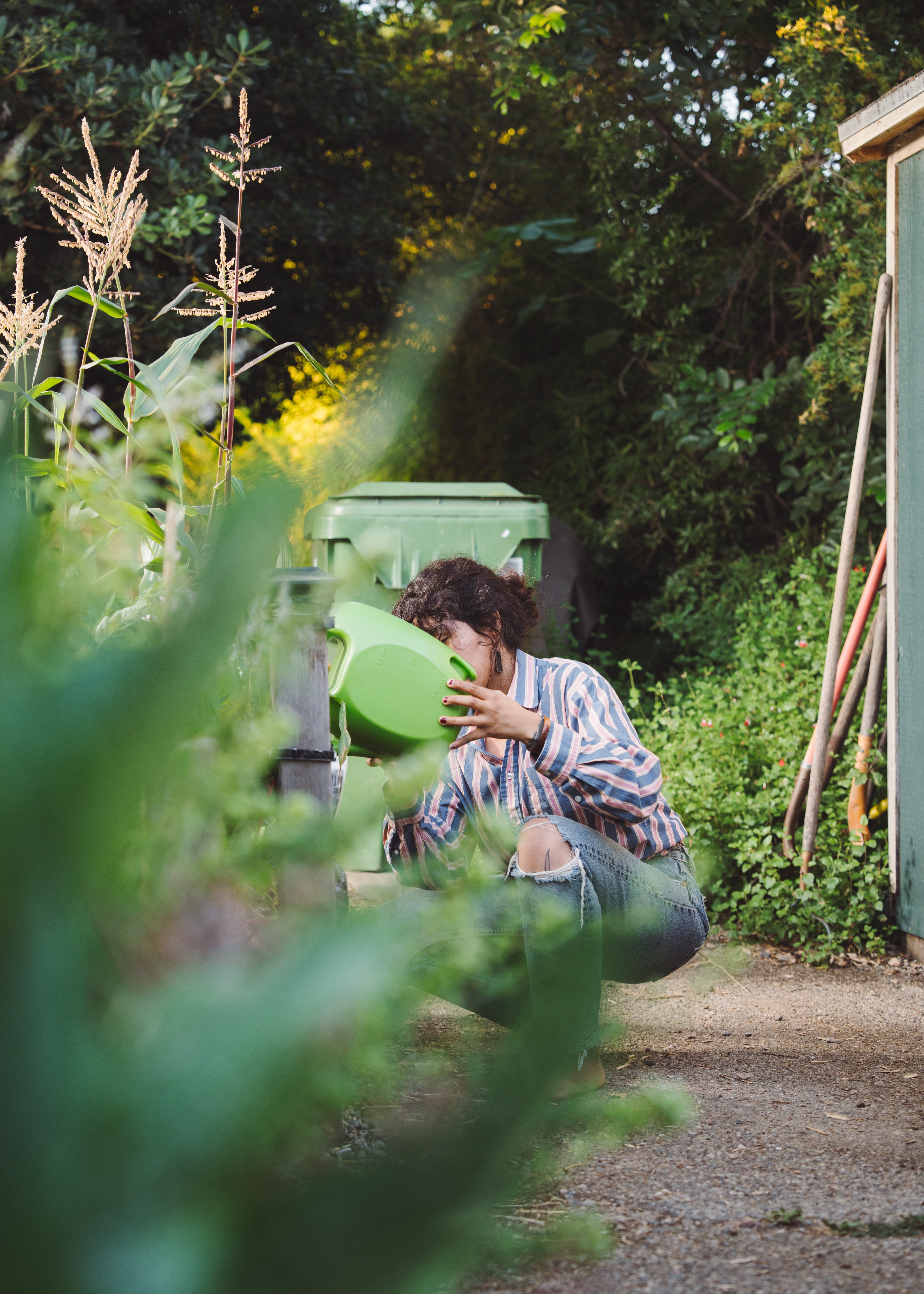 A woman squats in a garden, watering plants. Her face is obscured behind a green plastic watering can.
