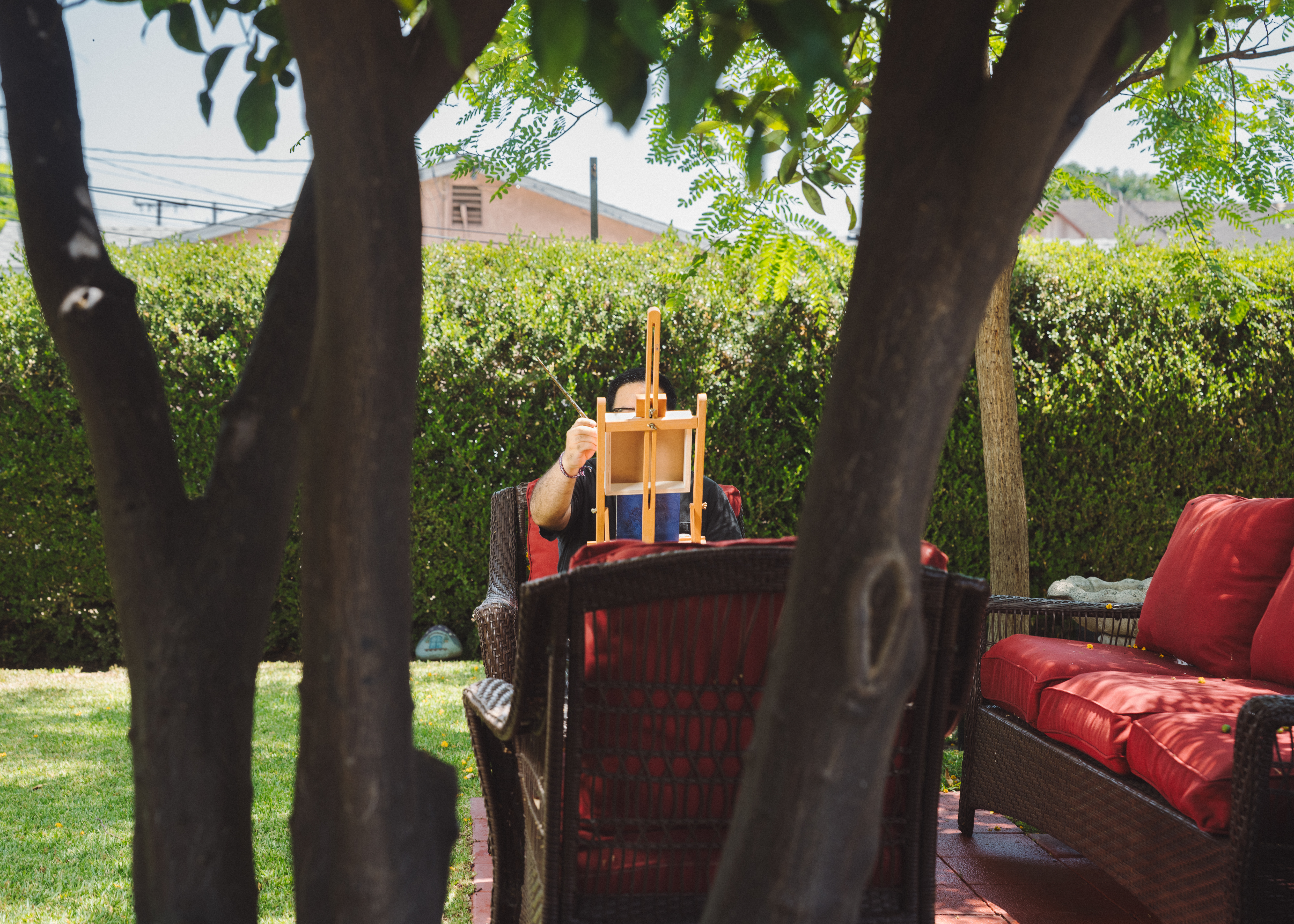 A man sits in a chair outside on a shaded patio, his face hidden behind a small easel.