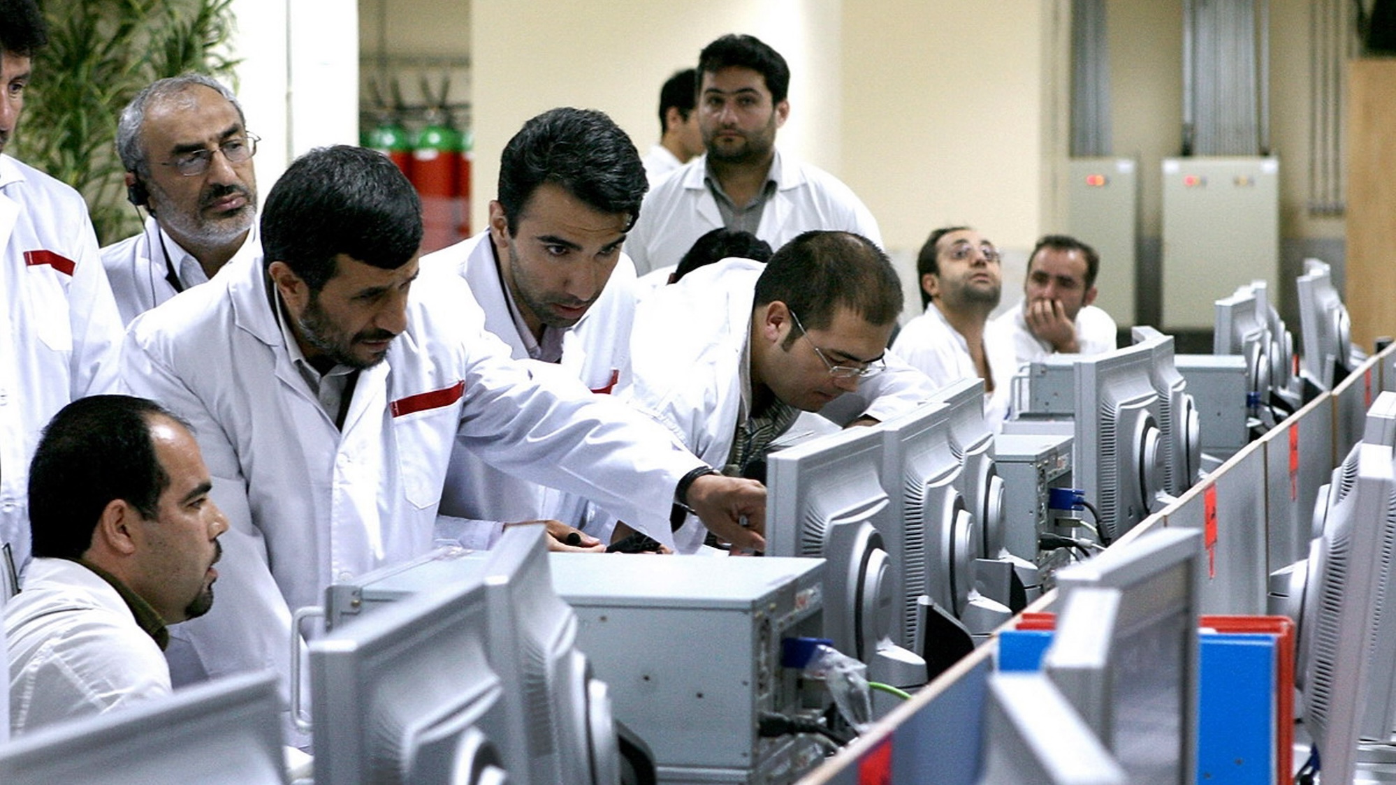 Several men wearing white shirts, crowd around screens and equipment.