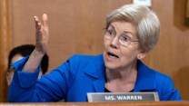Elizabeth Warren, who wears an electric blue blazer and sits behind a name card that reads her name, speaks animatedly with her hand gestured to person off camera.