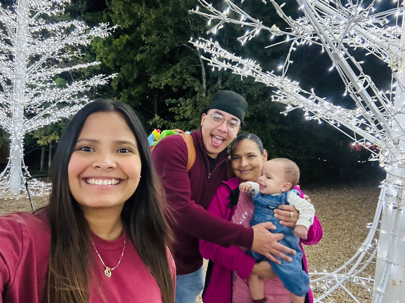A woman standing amid white metal trees with Christmas lights takes a photo while a man and older woman holding a baby pose behind her
