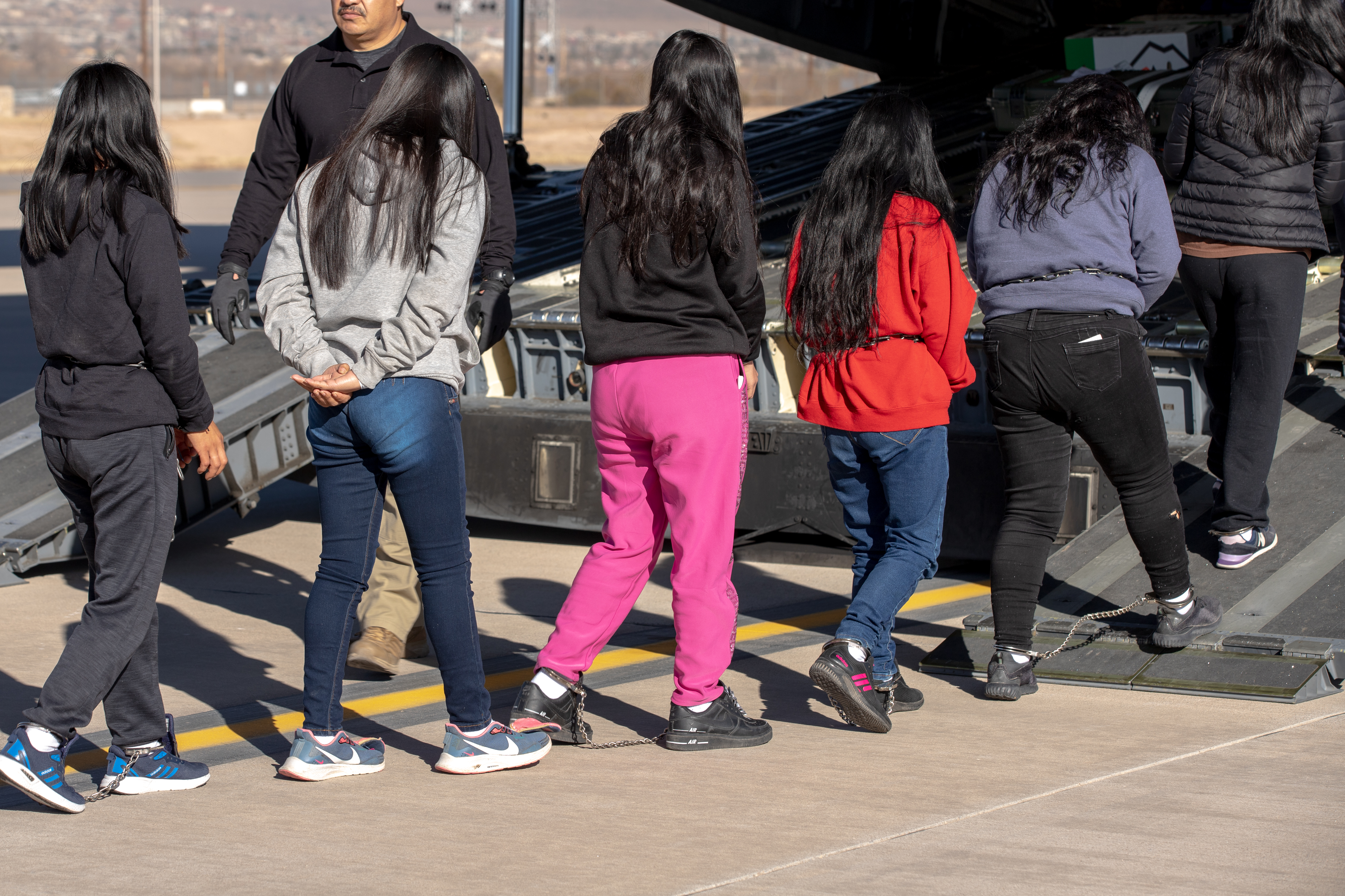 Six women, some with chains around their ankles and wrists, are escorted onto a military plane.