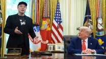Elon Musk standing, wearing a black jacket over a graphic t-shirt and a baseball cap, next to a seated Donald Trump, wearing a navy suit and red striped tie, in front of a row of flags in the Oval Office.