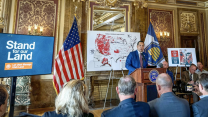 A man stands at a podium in front of a golden background and a map of the US with red markings.