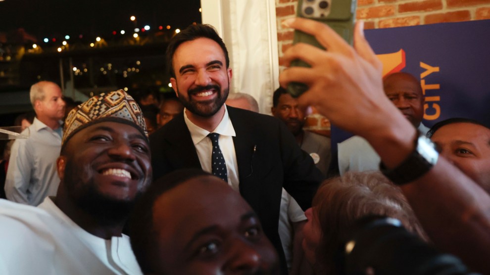 Zohran Mamdani in a suit, smiling and taking a selfie with a group of supporters