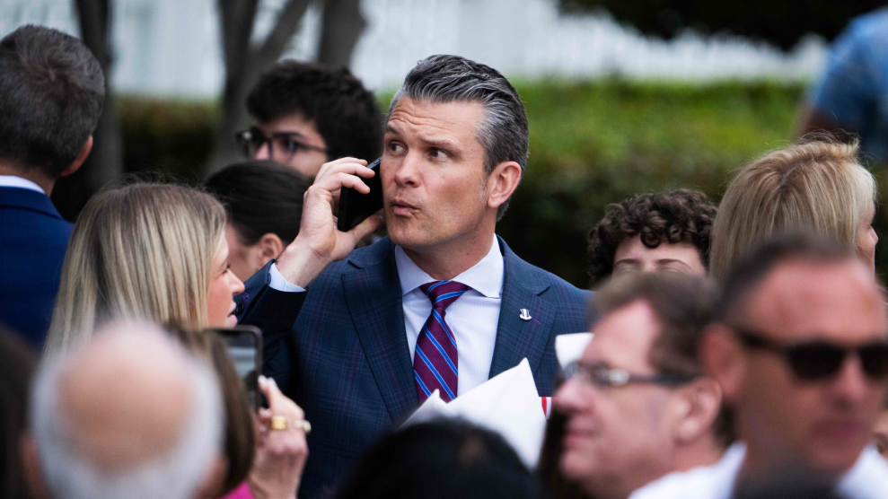 A middled-aged Caucasian man with slicked-back hair, standing amid a crowd of people, holds a mobile phone up to his ear. He is wearing a suit and tie and has a look of slight concern on his face.