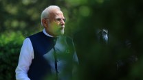 Indian Prime Minister Narendra Modi looks off to the side as he stands in a garden. The shot is partially obscured by out-of-focus leaves in the foreground.