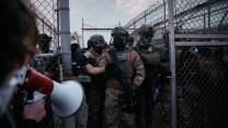 A protestor speaks into a megaphone, facing a line of armed law enforcement officers in tactical gear standing behind a chain-link fence.