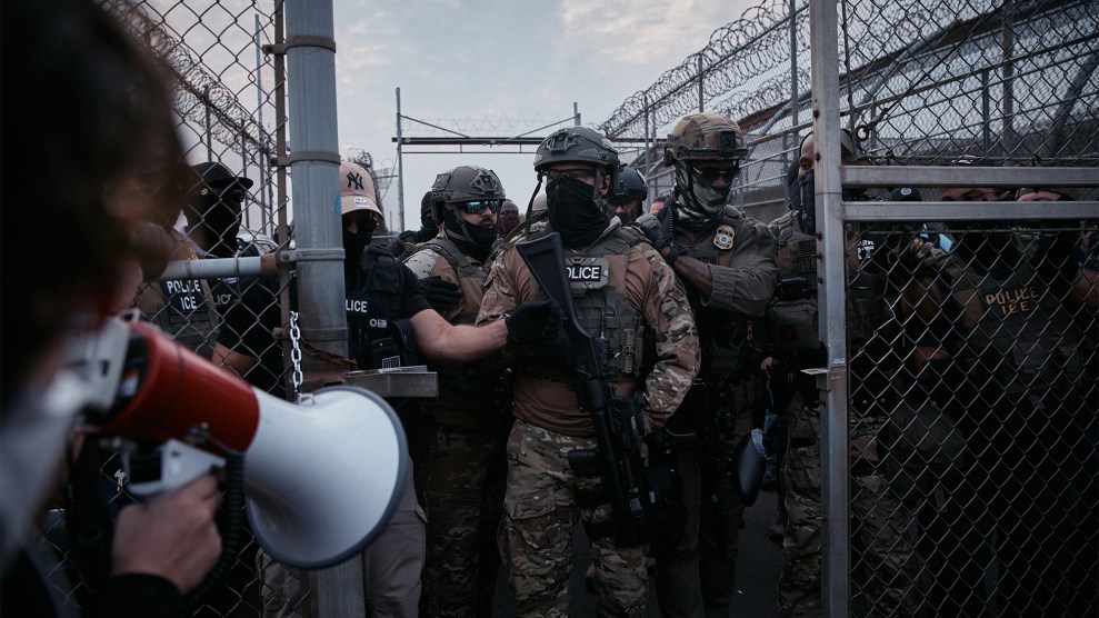 A protestor speaks into a megaphone, facing a line of armed law enforcement officers in tactical gear standing behind a chain-link fence.