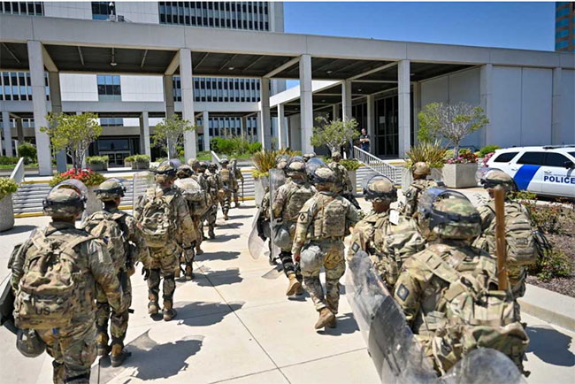 Two rows of national guard troops in full fatigues and helmets walks away from the camera on a sunny day and toward the staircase of a federal building in los angeles