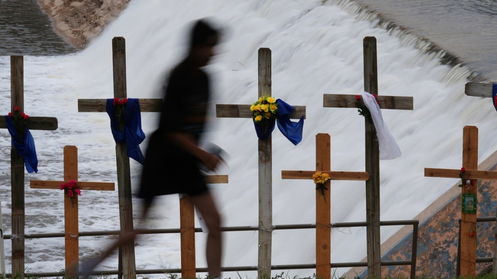 A blurred figure walks past a row of wooden crosses placed along a railing in front of a rushing dam spillway. The crosses are adorned with colorful flowers, blue and white fabric, and other memorial decorations, indicating a tribute or memorial site. The scene conveys a somber and reflective mood.