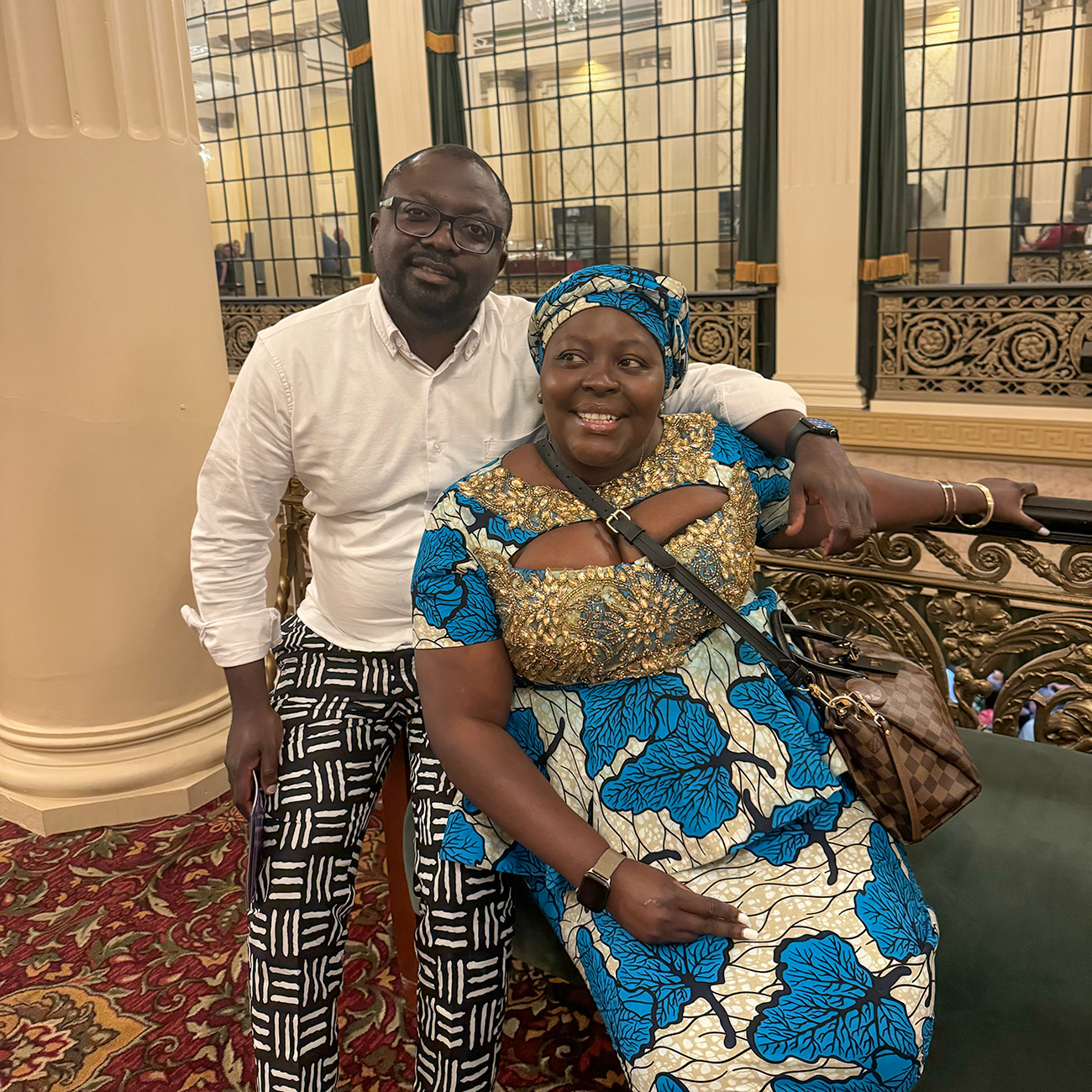 A man in a white shirt and patterned pants stands with his arm around a woman in a vibrant blue and gold patterned dress and matching headwrap.