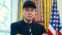 Picture of Musk in the Oval Office wearing a black DOGE baseball cap.