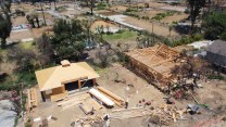 Aerial image primarily of two plots of land with homes in different stages of construction. There are piles of wood in front of the homes, and beyond them scorched earth without buildings.