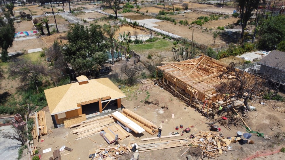 Aerial image primarily of two plots of land with homes in different stages of construction. There are piles of wood in front of the homes, and beyond them scorched earth without buildings.