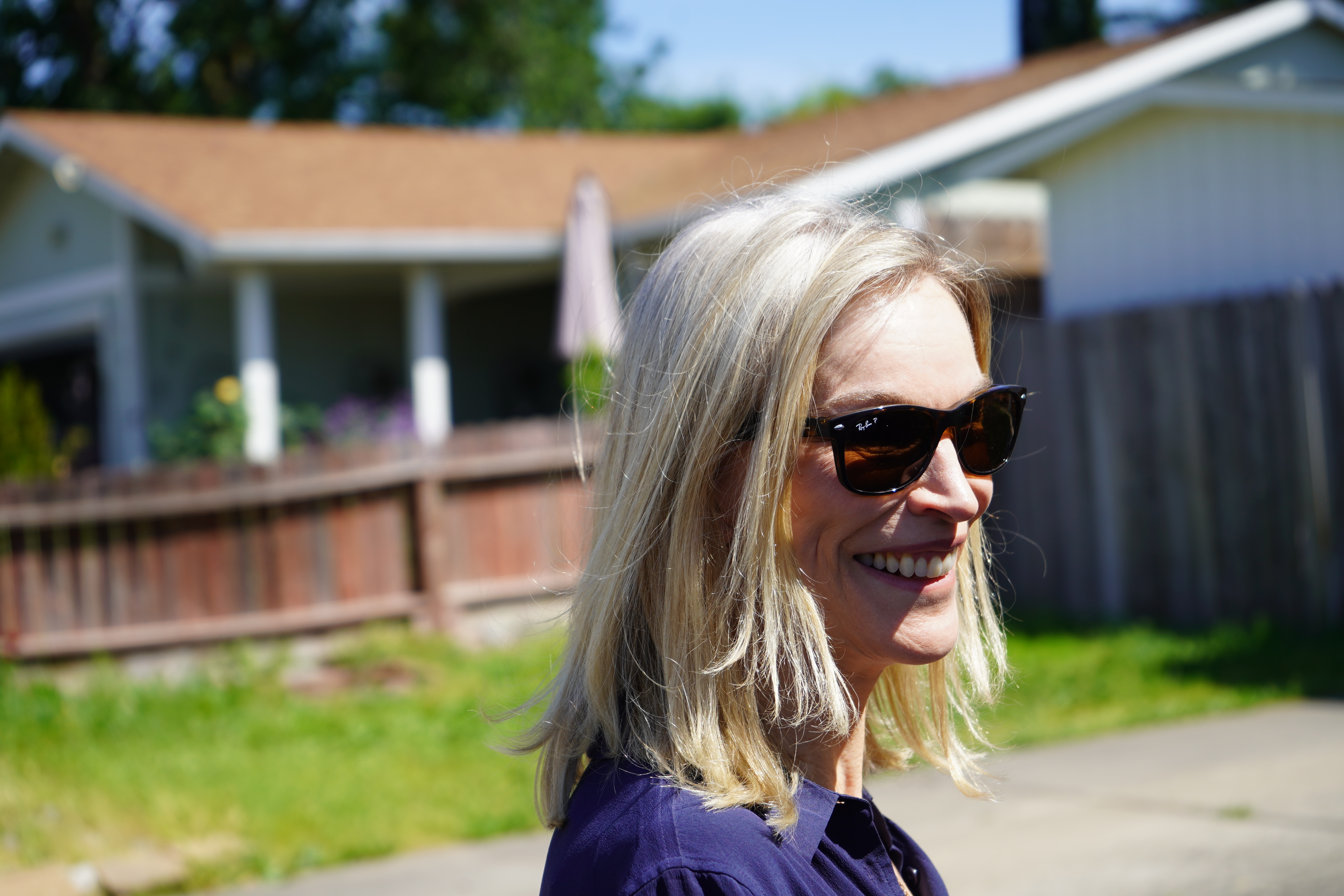 A blond woman in sunglasses stands outside a house with a brown wooden fence.