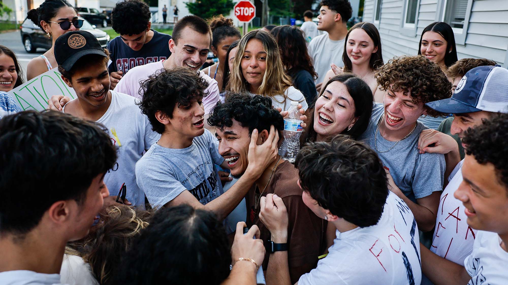 A smiling boy in a brown shirt is surrounded and hugged by a group of people wearing white and grey shirts.