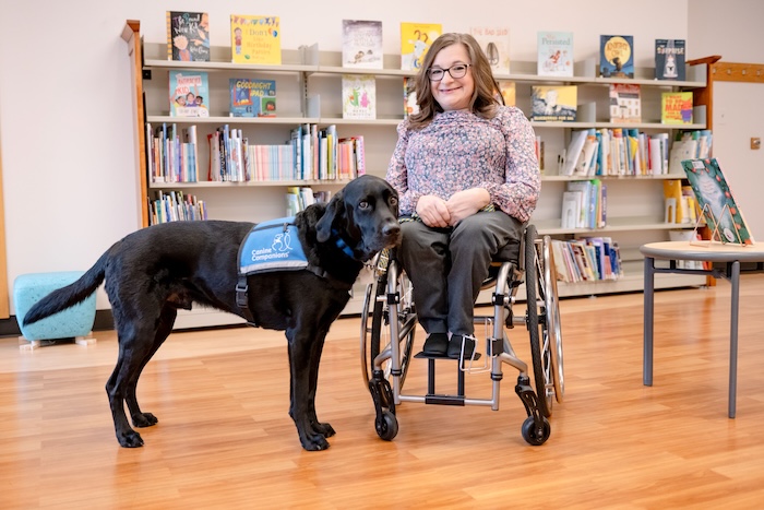 A white woman in a wheelchair with a black service dog in a library