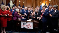 A group of over a dozen people dressed in suits or pantsuits. They clap and shake hands. Mike Johnson sits at a desk in the center of them. On the desk is a sign that says "One Big Beautiful Bill Act."